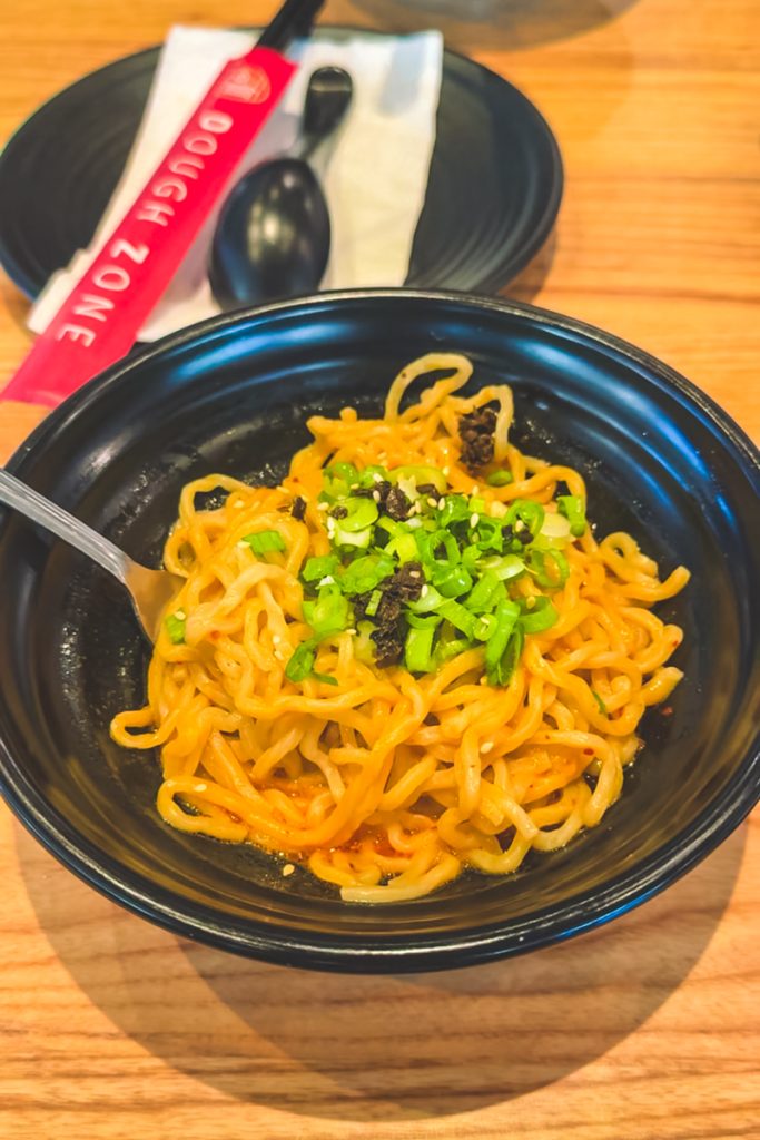 Bowl of dan dan noodles topped with chopped green onions served on a wooden table with chopsticks and spoon