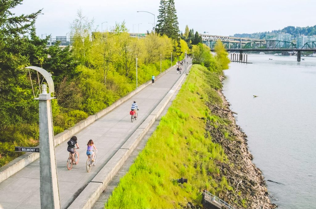 People biking and walking along a paved riverfront path beside calm water with trees and bridges in the distance