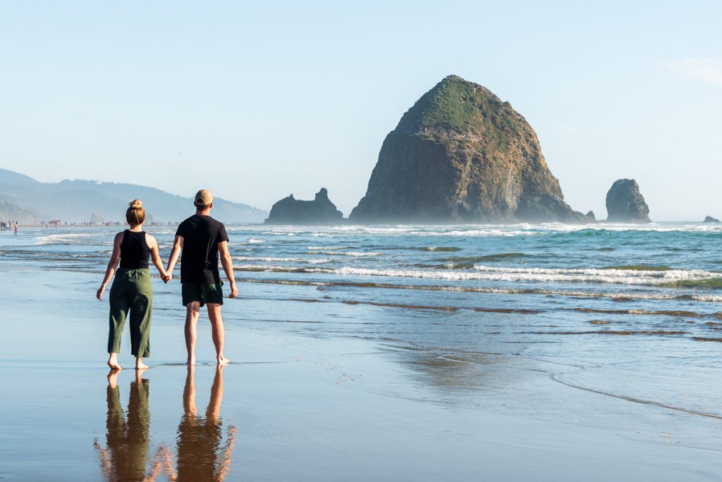 A couple walking barefoot along the shoreline at Cannon Beach with Haystack Rock rising from the ocean, one of the iconic things to see near Portland