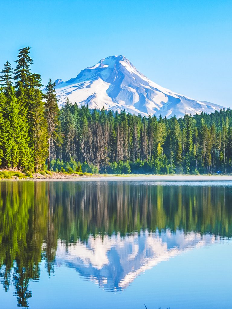 A calm lake reflecting snow-capped Mount Hood and evergreen forest under a bright blue sky, highlighting one of the most scenic things to see near Portland