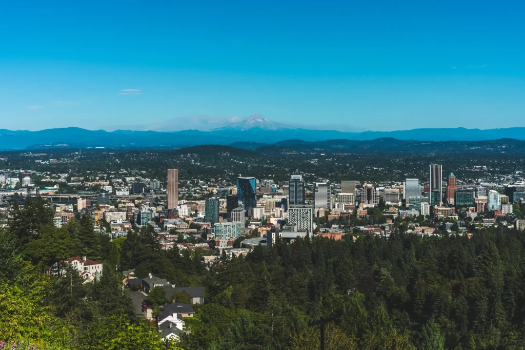 Panoramic view of the downtown Portland, Oregon skyline from Pittock Mansion, with Mount Hood rising in the distance and dense evergreen forests surrounding the city.