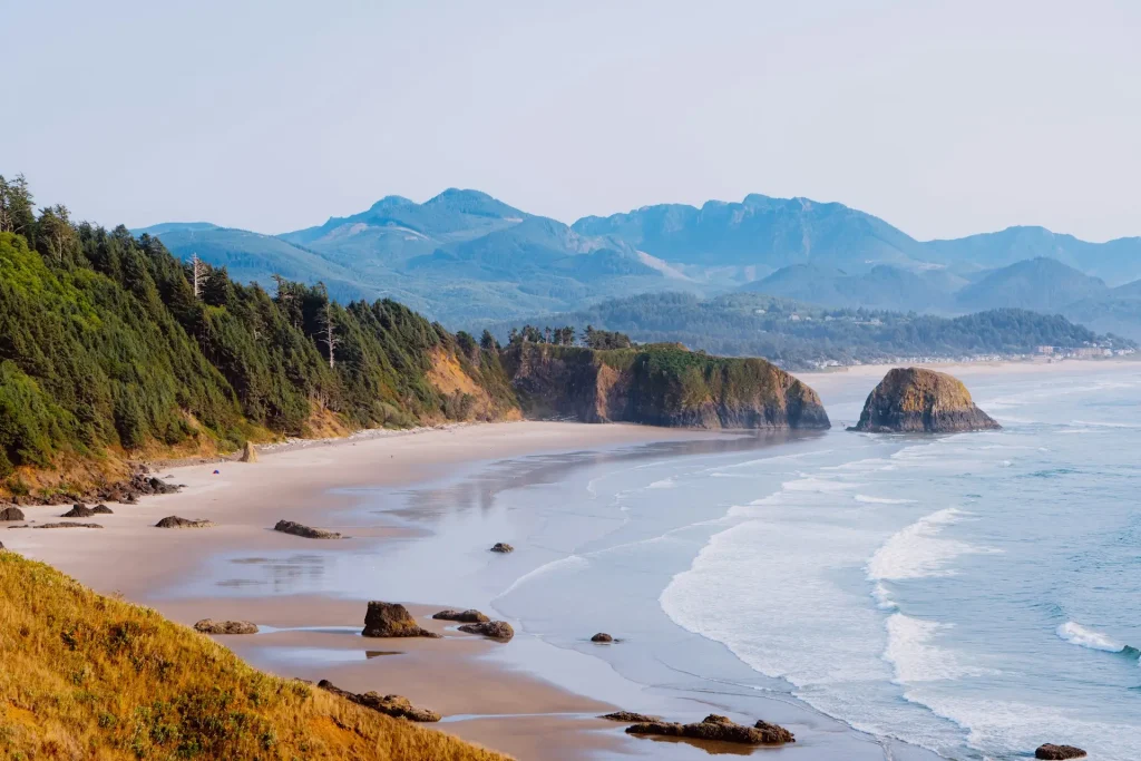 Scenic coastal view from Ecola State Park in Oregon, featuring a sandy beach, rocky sea stacks, forested cliffs, and mountains along the Pacific Ocean.