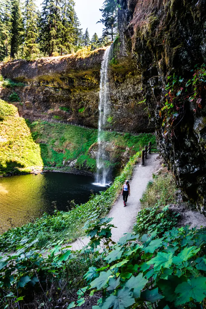 Hiker walking along the trail behind South Falls at Silver Falls State Park in Oregon, with the waterfall plunging over a rocky cliff into a forested pool.