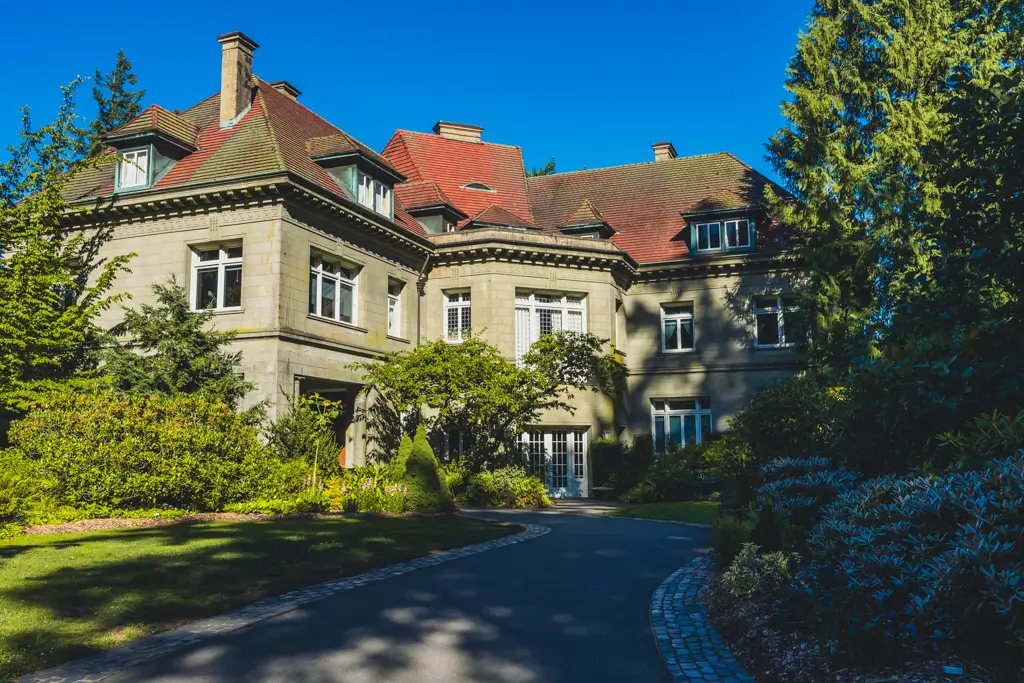 Exterior of the historic Pittock Mansion in Portland, Oregon, surrounded by lush green gardens and tall trees beneath a bright blue sky.
