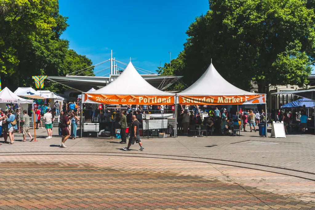 Entrance to the Portland Saturday Market in Portland, Oregon, with white vendor tents, crowds browsing local booths, and large green trees on a sunny day.