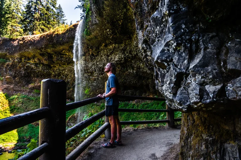 JT Mocarski of The Mobile Homie standing along the trail behind South Falls at Silver Falls State Park in Oregon, surrounded by basalt cliffs and lush forest.