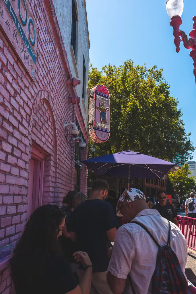 Visitors lined up outside the original Voodoo Doughnut in Portland, Oregon, beside its pink brick exterior and colorful neon sign on a sunny day.