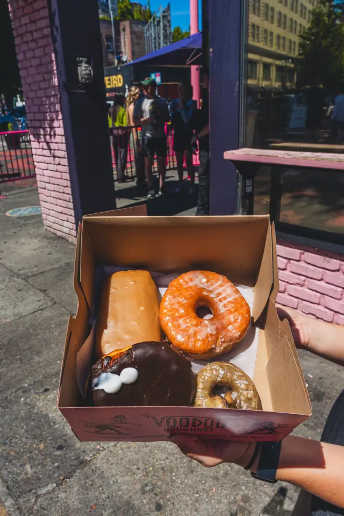 Open box of four colorful Voodoo Doughnuts held outside the iconic pink Voodoo Doughnut shop in downtown Portland, Oregon.