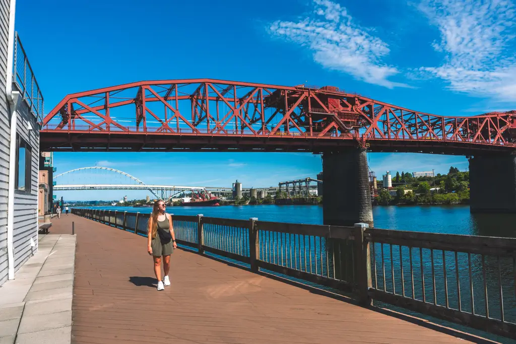 Woman walking along the waterfront in Portland, Oregon, beneath the red Steel Bridge with the Willamette River and Fremont Bridge visible in the distance.