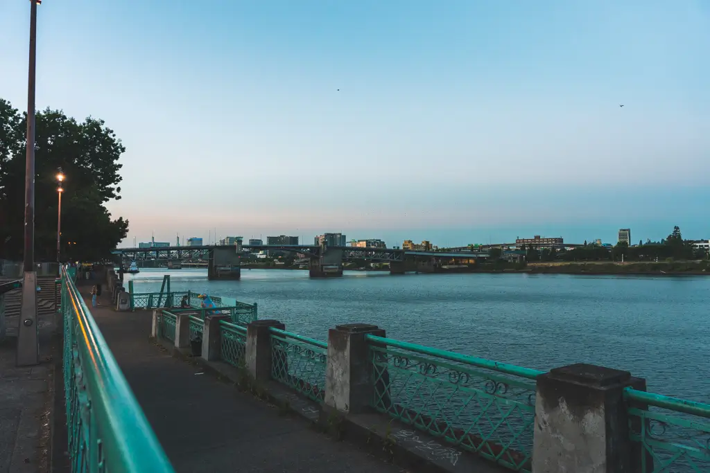 Portland waterfront path along the Willamette River at dusk, with green railings, city bridges, and the Portland skyline visible across the water.