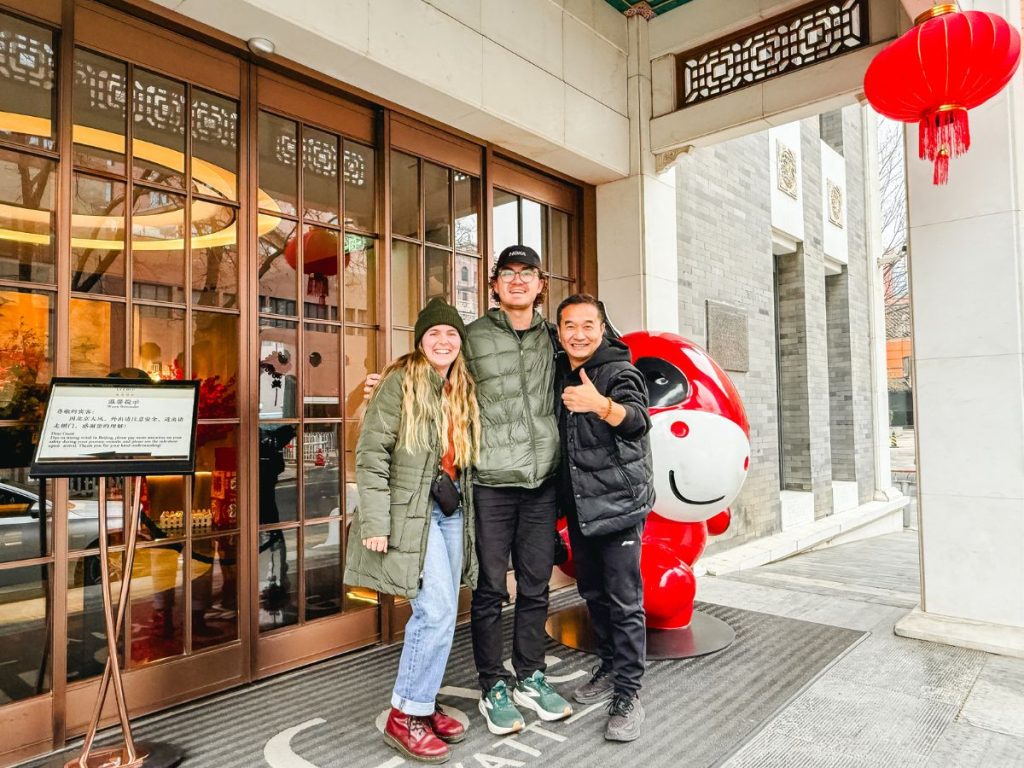 Three people smiling outside a traditional-style building in Beijing, with red lanterns overhead and a large cartoon statue beside them, posing together on a city street.
