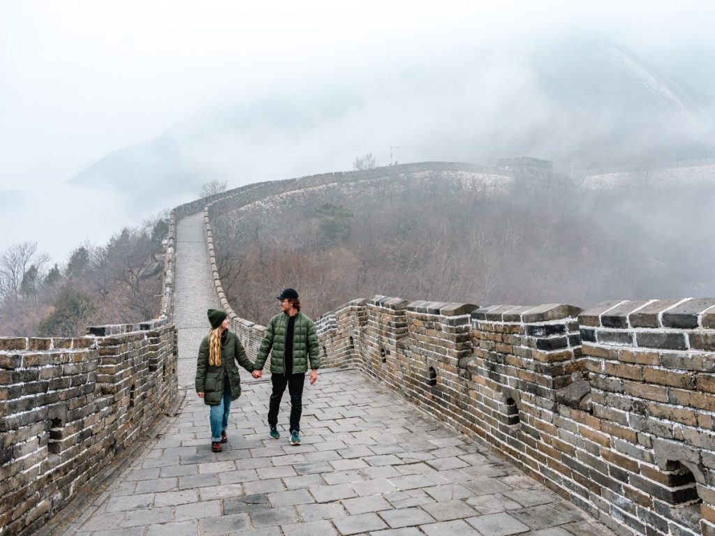 Couple holding hands while walking along the Great Wall of China on a misty day, surrounded by stone walls and soft fog covering the mountains in the background.