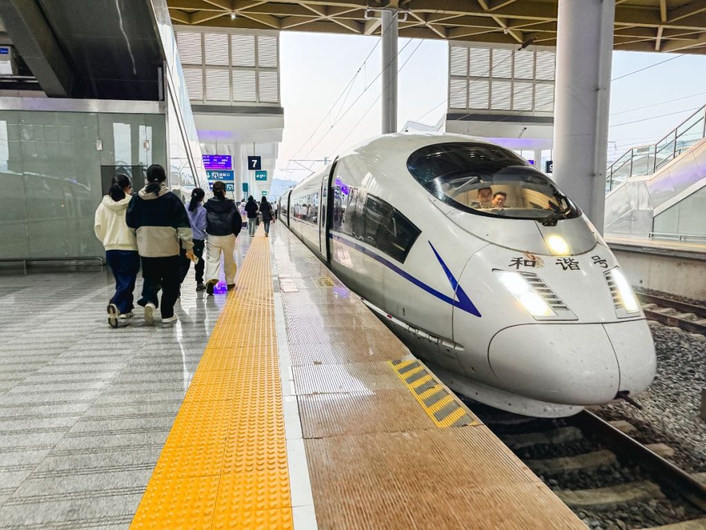 Modern high-speed train stopped at Zhangjiajie railway station platform with passengers walking alongside, featuring sleek design and bright indoor station lighting.