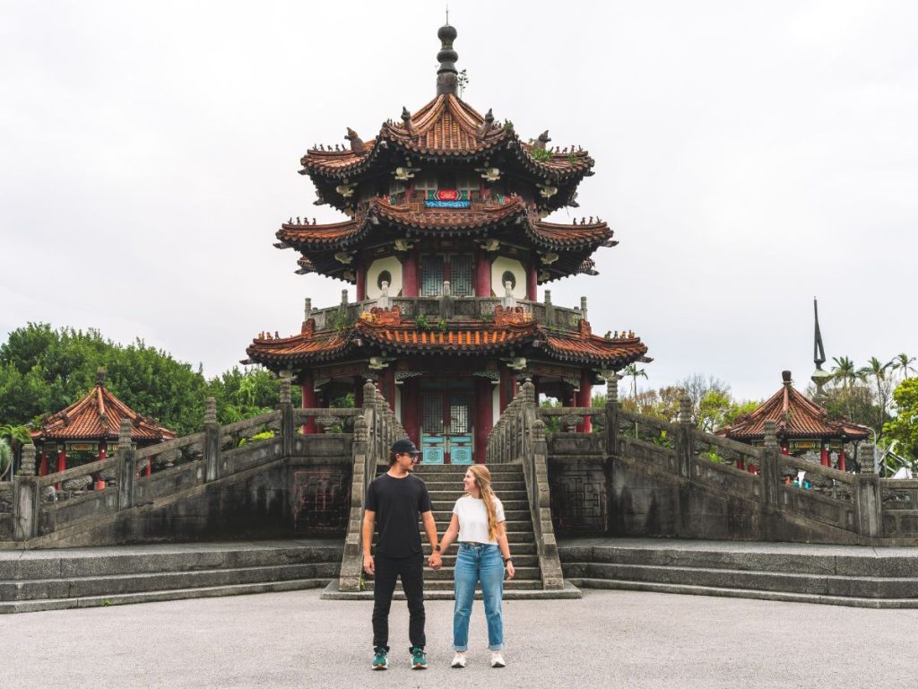 Couple holding hands in front of an ornate multi-tiered traditional Chinese pavilion with curved roofs, stone steps, and detailed architecture in a peaceful outdoor setting in Taiwan.