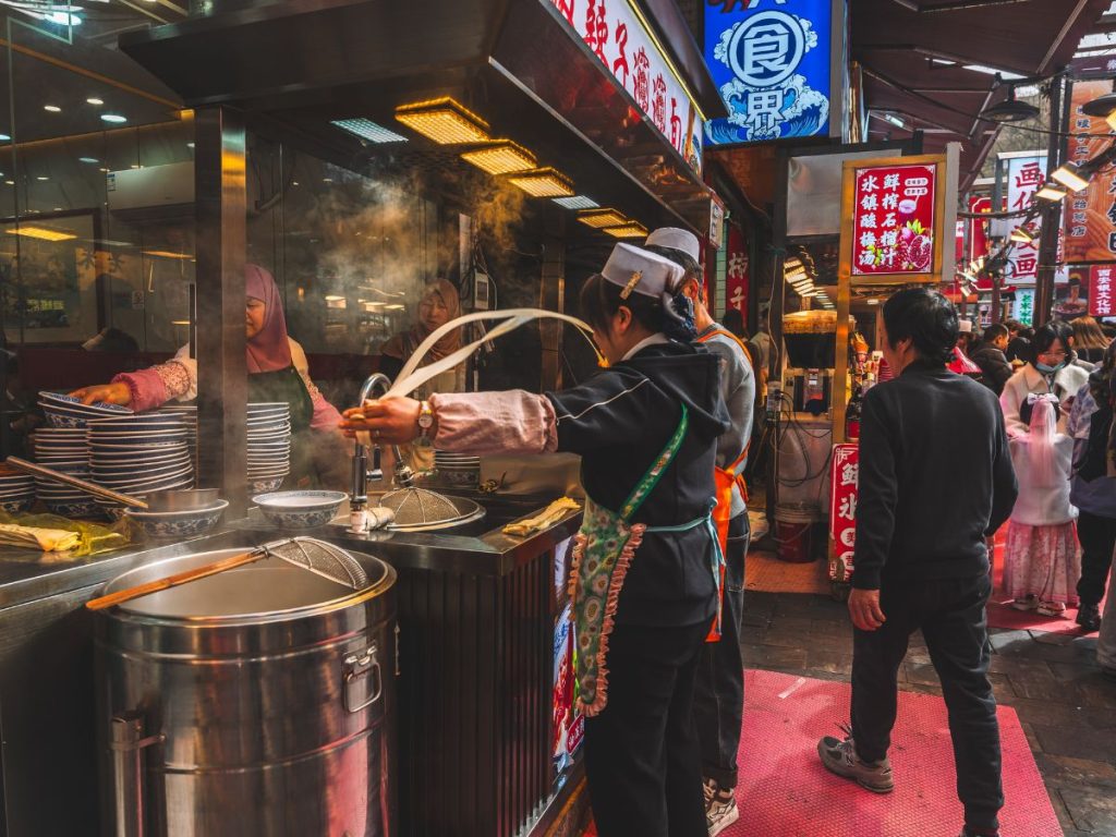 Street vendor preparing fresh noodles at a busy outdoor food stall in Xi’an’s Muslim Quarter, with steam rising from pots and people walking through the vibrant market.