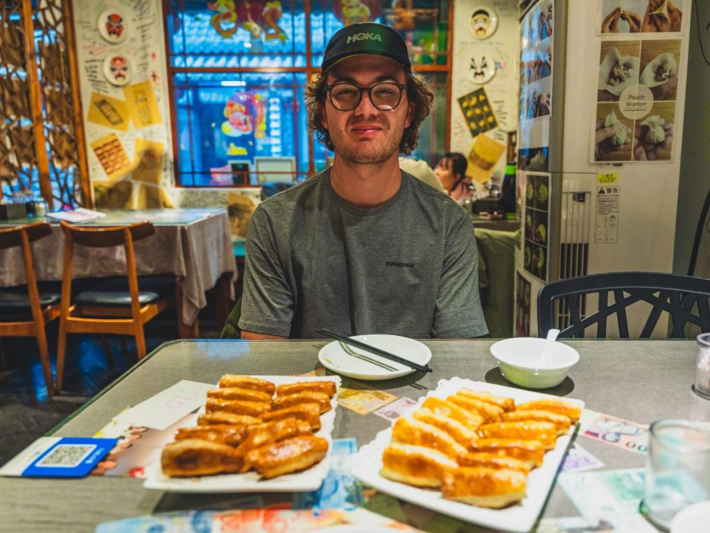 Man sitting at a table at Mr. Shi’s Dumplings in Beijing with plates of pan-fried dumplings in front of him, inside a cozy restaurant filled with colorful notes and decor on the walls.