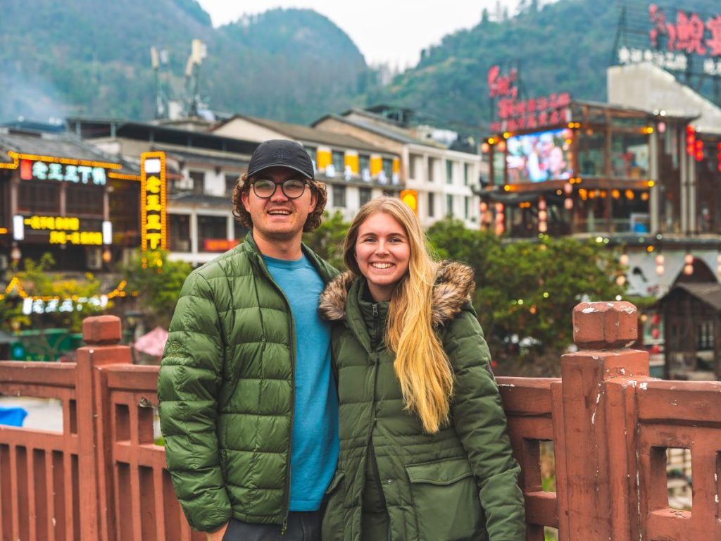 Couple standing on a bridge in Wulingyuan near Zhangjiajie, China, with colorful buildings, bright signage, and misty green mountains in the background.