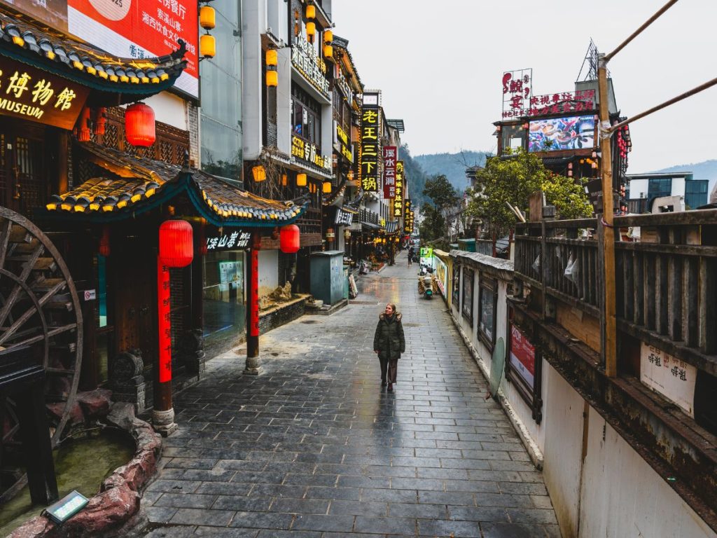 Person walking down a quiet street in Wulingyuan near Zhangjiajie, China, lined with traditional-style buildings, red lanterns, and glowing shop signs after rainfall.