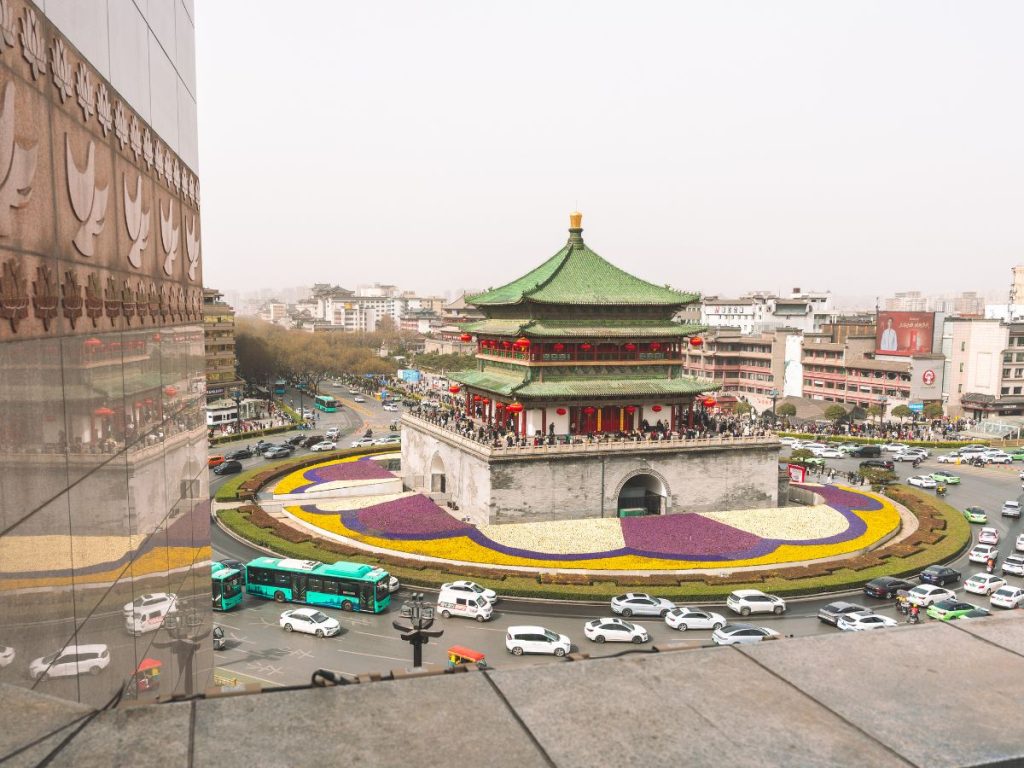 Aerial view of the Xi’an Bell Tower surrounded by a busy roundabout with cars, buses, and pedestrians, featuring traditional Chinese architecture with a green roof and red lanterns.