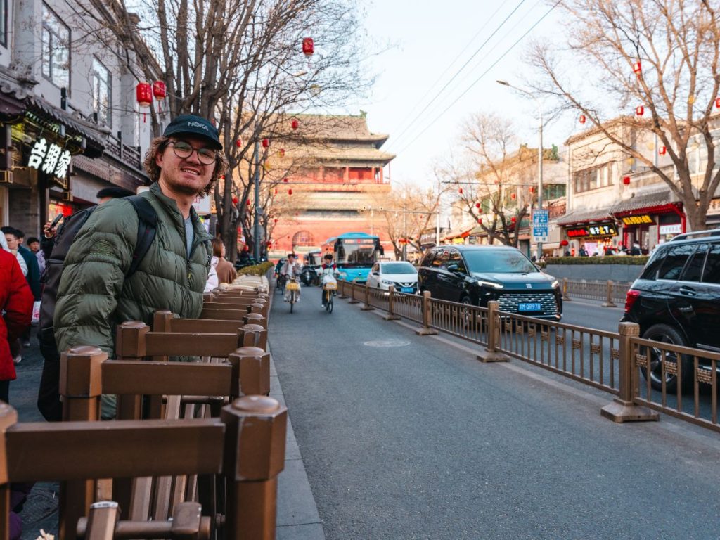 Man standing along a busy street in Beijing with red lanterns hanging overhead, as cars, bikes, and pedestrians move through the city with traditional Chinese architecture visible in the background.