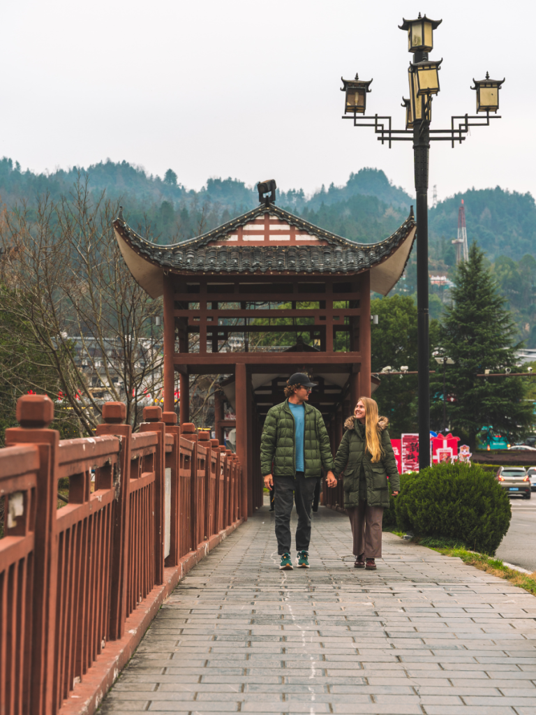 Couple holding hands while walking along a traditional-style bridge in Wulingyuan near Zhangjiajie, China, with wooden architecture, lantern-style street lamps, and misty green mountains in the background.