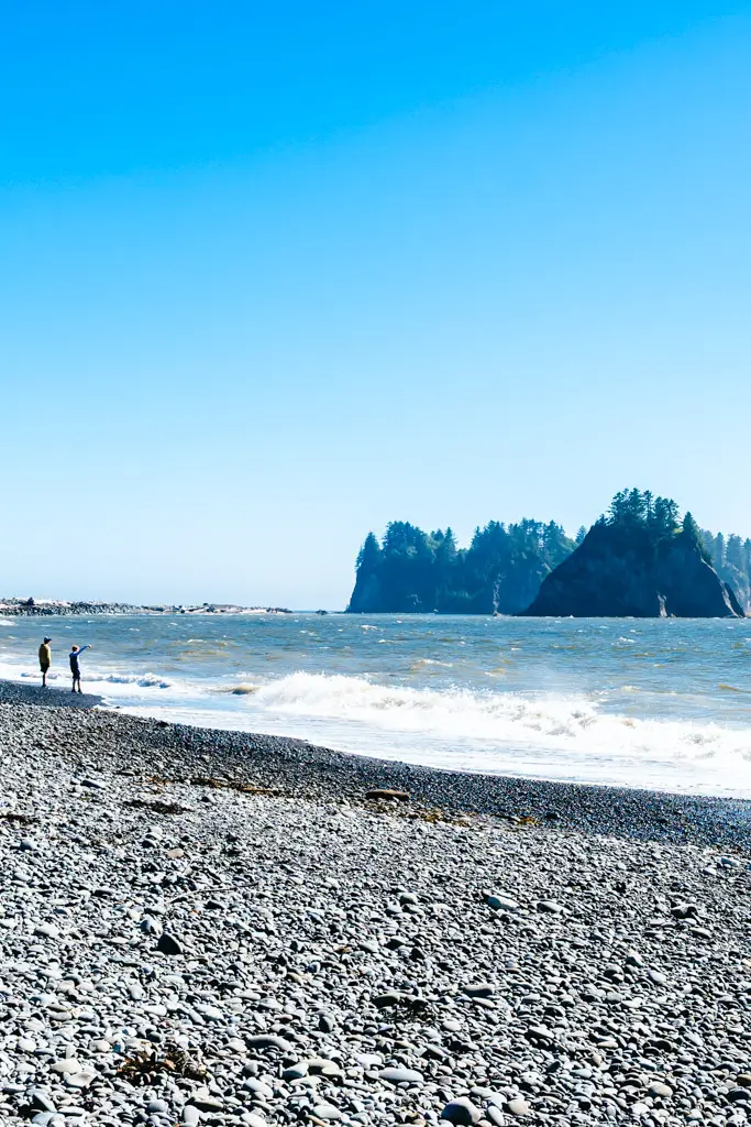 Rialto Beach in Olympic National Park with a rocky shoreline, crashing Pacific waves, and dramatic sea stacks rising from the ocean beneath a bright blue sky in Washington.