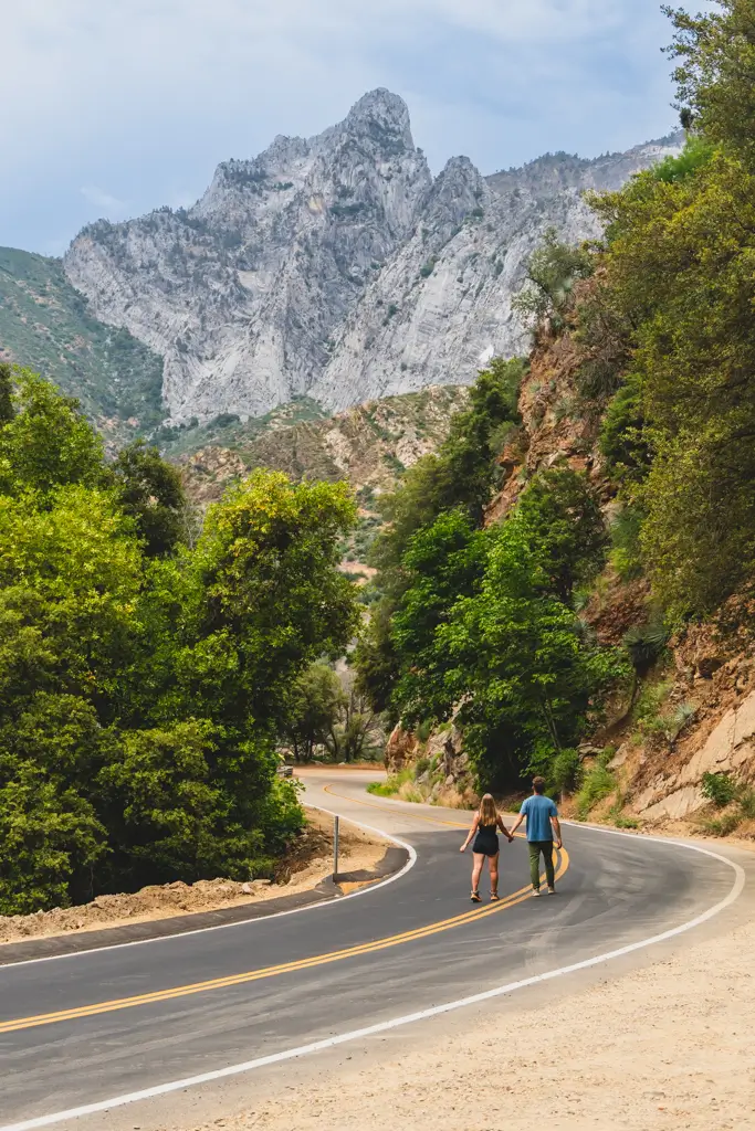 Couple walking along a winding mountain road surrounded by towering granite cliffs and lush forest in Kings Canyon National Park, California.