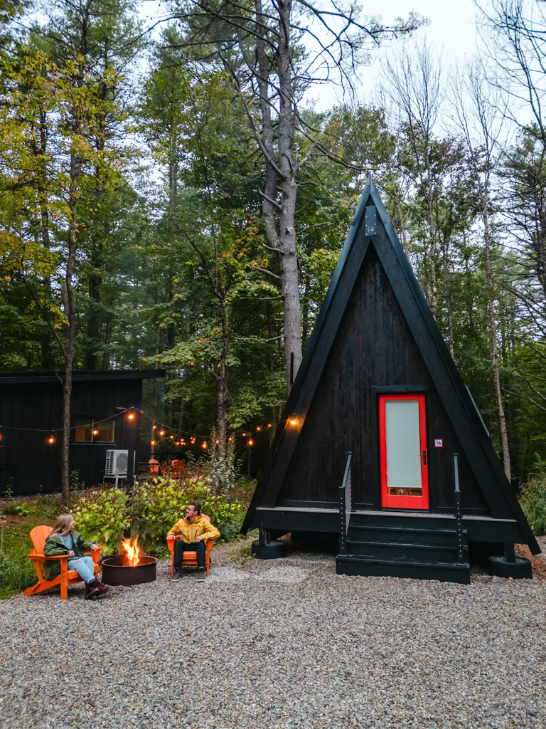 Black A-frame cabin with a bright red door at the Desert of Maine Campground in Freeport, Maine, with travelers relaxing around a campfire under string lights.