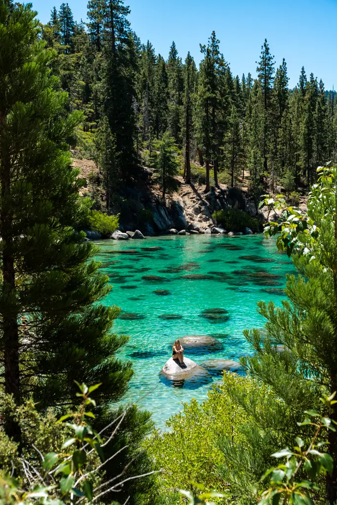 Traveler sitting on a boulder in the crystal-clear turquoise waters of Bonsai Rock at Lake Tahoe, surrounded by granite rocks and towering pine trees.
