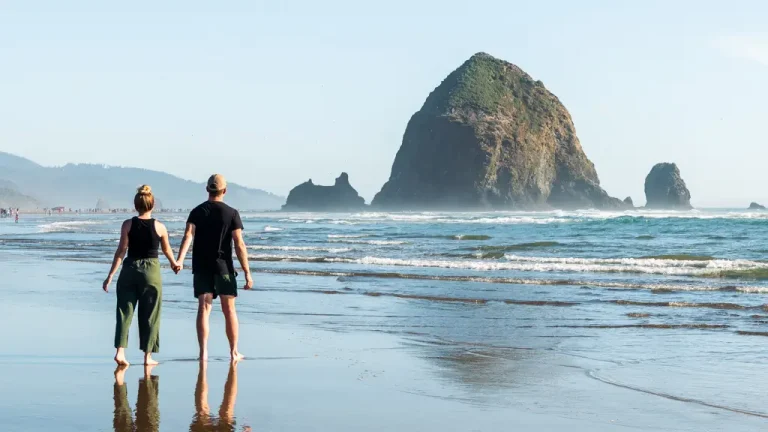 Couple holding hands on Cannon Beach with Haystack Rock, one of the Pacific Northwest's most iconic coastal landmarks, on a sunny day along the Oregon Coast.
