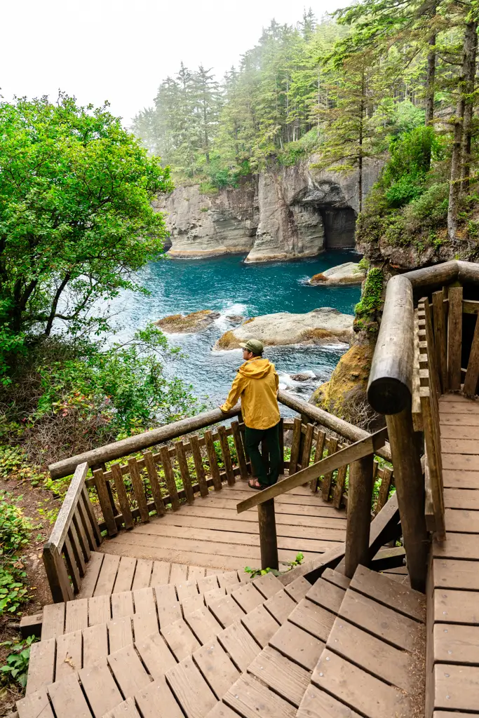 Traveler standing on a wooden viewing platform overlooking the sea caves and turquoise waters at Cape Flattery, Washington, surrounded by lush coastal forest.