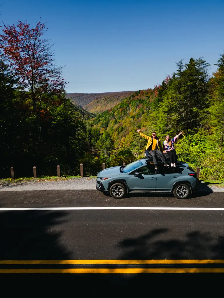 Couple sitting on the roof of a Subaru overlooking a scenic mountain road surrounded by colorful fall foliage in Blackwater Falls State Park, West Virginia.