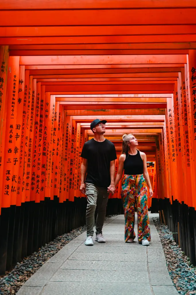 Couple walking hand in hand beneath the iconic red torii gates at Fushimi Inari Taisha in Kyoto, Japan, looking up at the historic shrine pathway.