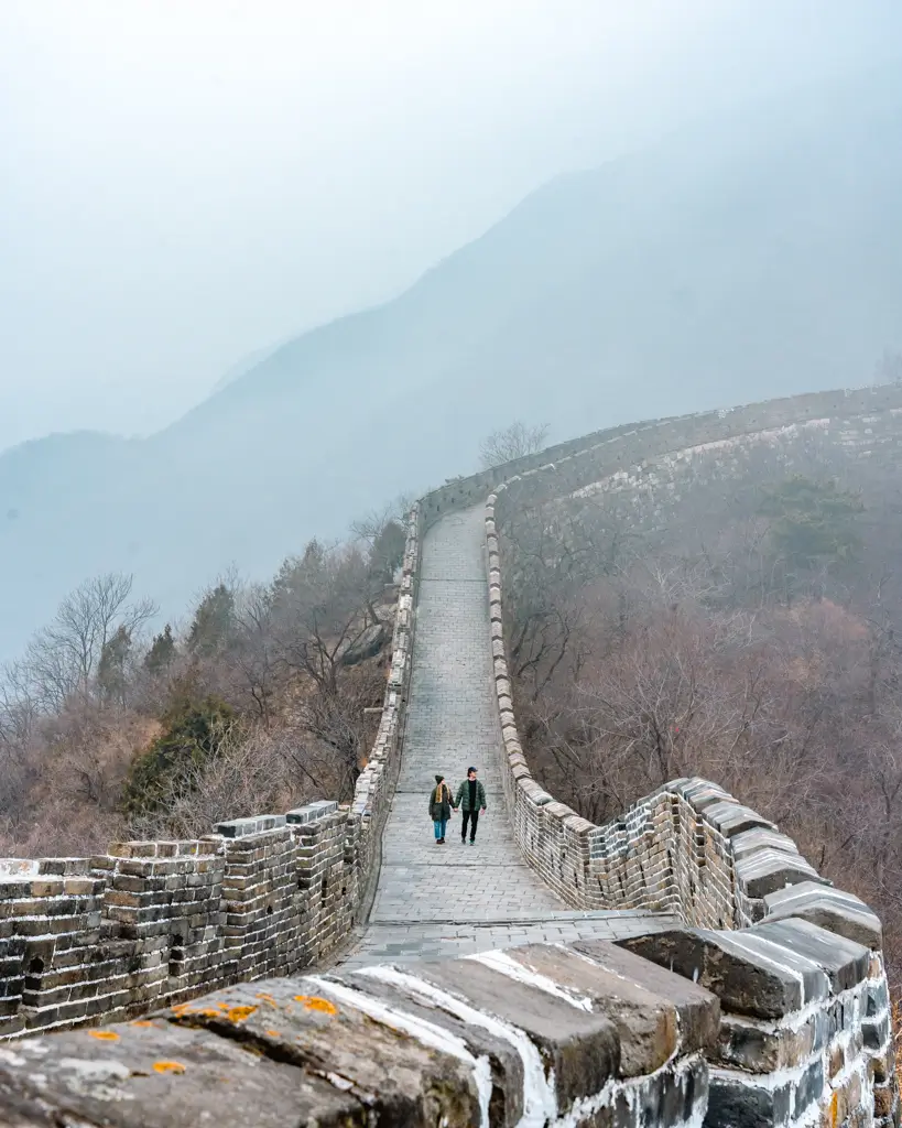 Two travelers walking along the historic stone pathway of the Great Wall of China at Mutianyu, surrounded by misty mountains and winter scenery.