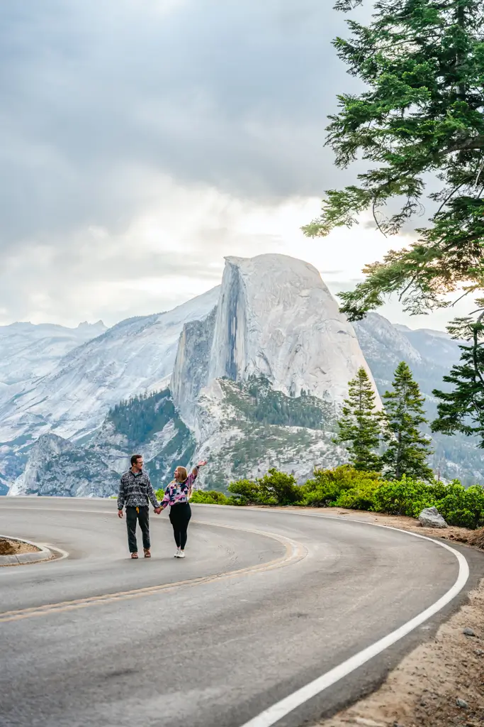 Couple walking hand in hand along Glacier Point Road with a stunning view of Half Dome in Yosemite National Park, California.