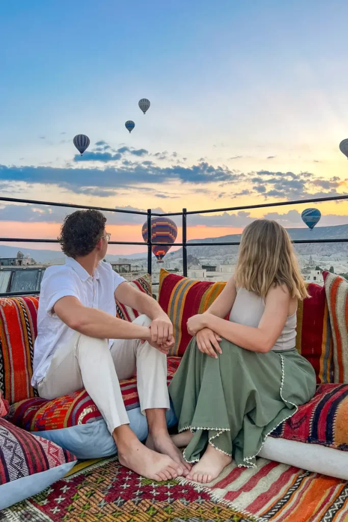 Couple sitting on a rooftop terrace watching colorful hot air balloons float over Cappadocia, Turkey, during sunrise.