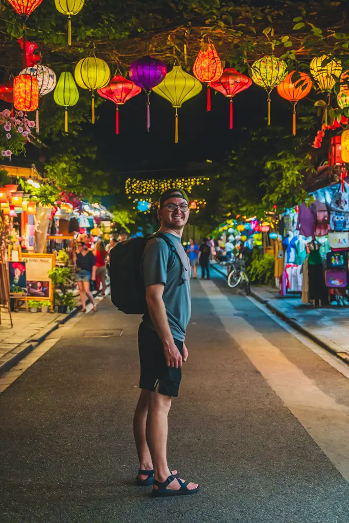 Traveler standing beneath colorful lanterns in the illuminated streets of Hoi An Ancient Town, Vietnam, surrounded by shops and vibrant night market lights.