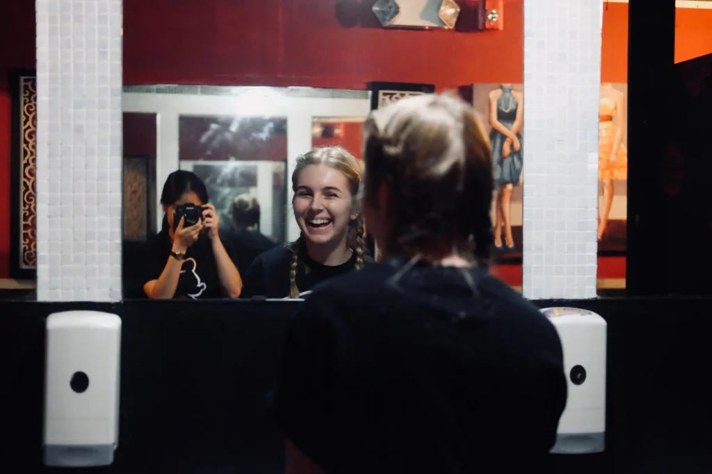Traveler laughing in front of a restroom mirror while a friend takes a photo in the reflection, capturing a candid travel moment.