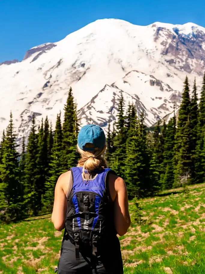 A hiker overlooking the snow-covered summit of Mount Rainier from the Sourdough Ridge Trail, surrounded by alpine meadows and evergreen trees in the Sunrise area of Mount Rainier National Park, Washington.