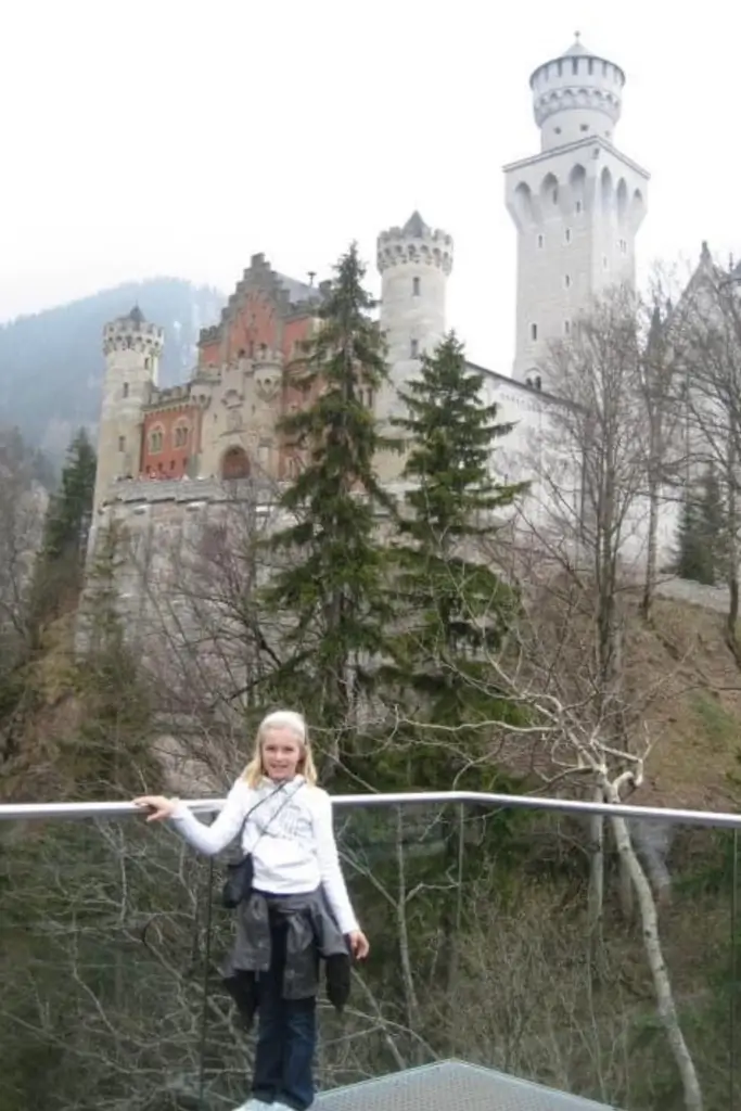 Young traveler standing at the Marienbrücke viewpoint overlooking Neuschwanstein Castle in Bavaria, Germany, with the fairytale castle rising above the surrounding forest.