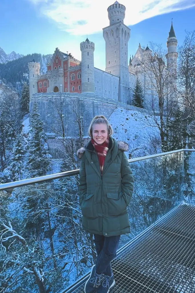 Traveler standing at the Marienbrücke viewpoint overlooking the snow-covered Neuschwanstein Castle in Bavaria, Germany, on a winter day.