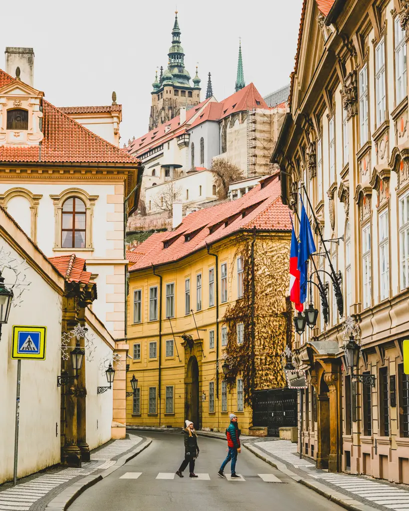 Two travelers crossing a quiet street in Prague's Castle District with the towers of Prague Castle rising above colorful historic buildings in the Czech Republic.