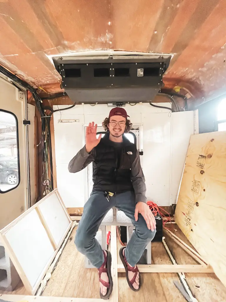 Traveler waving from inside a partially converted shuttle bus, showing exposed walls, flooring, and framing during the early stages of a DIY camper build.