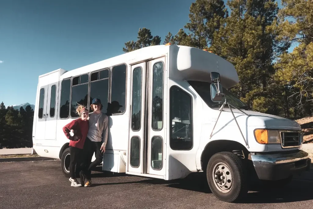 Couple posing beside a white shuttle bus surrounded by pine trees in Grand Canyon National Park, Arizona, during a scenic road trip.