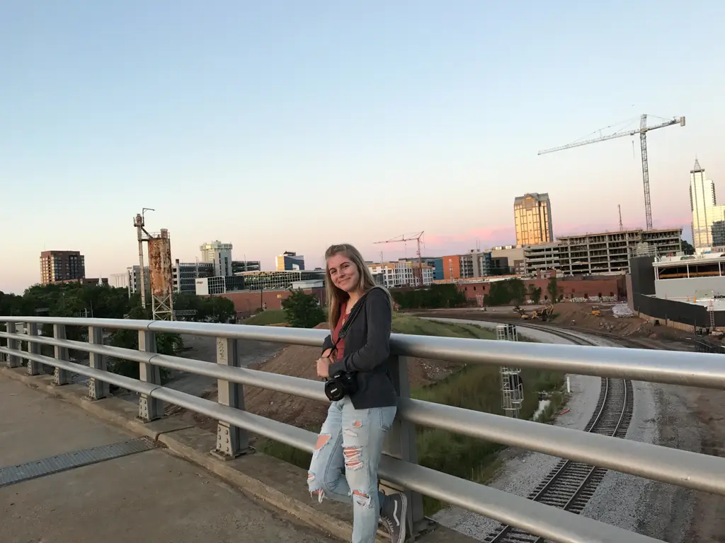 Traveler leaning against a bridge railing at sunset overlooking the Raleigh skyline, railroad tracks, and downtown buildings in North Carolina.