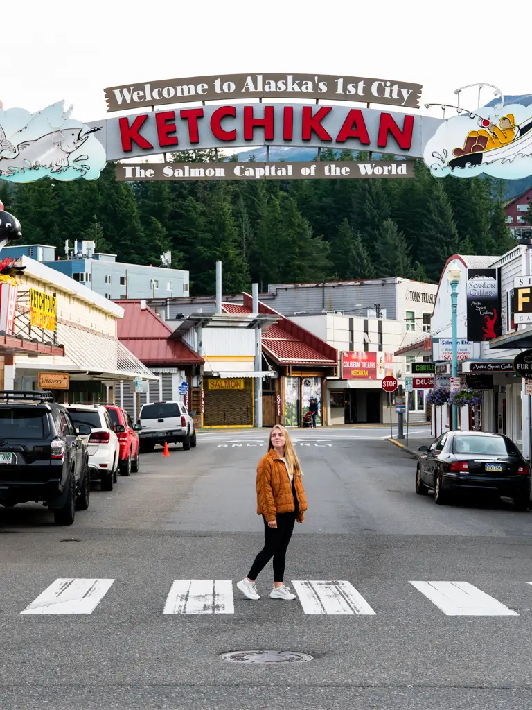 Traveler standing beneath the iconic "Welcome to Alaska's 1st City" sign in downtown Ketchikan, Alaska, known as the Salmon Capital of the World.