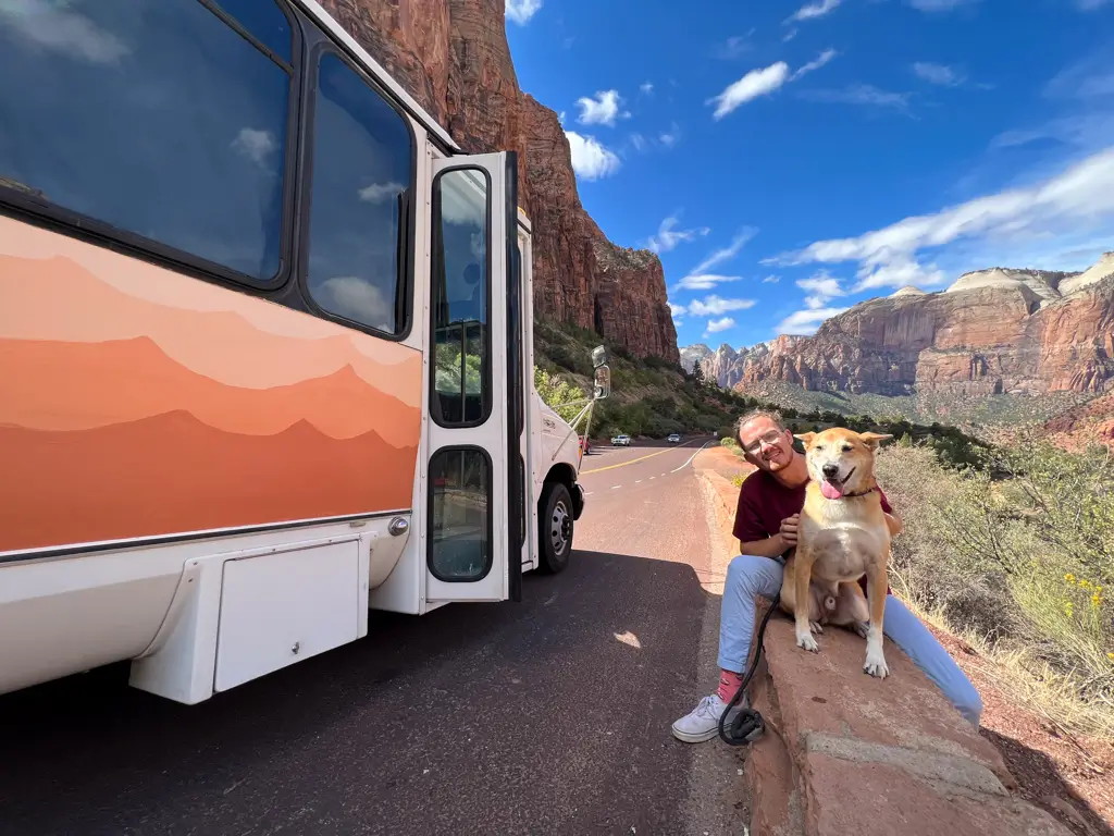 Traveler and dog sitting beside the Zion Canyon Shuttle with towering red sandstone cliffs in the background at Zion National Park, Utah.