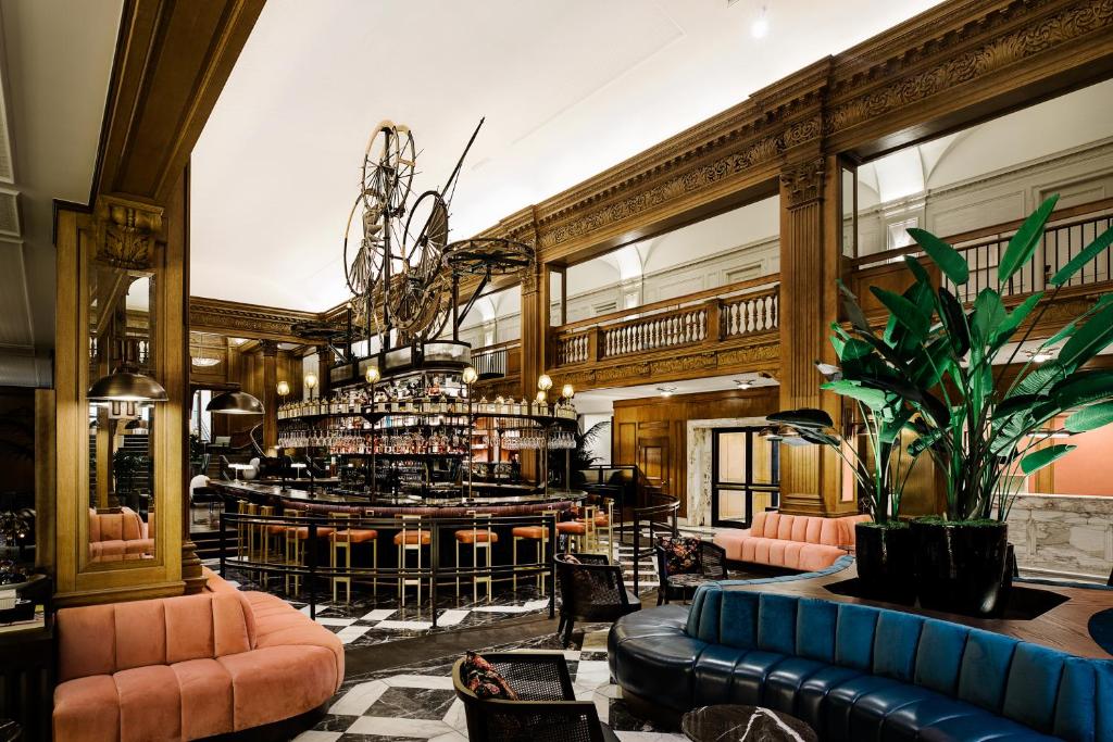 A grand hotel bar at the Fairmont Olympic Hotel featuring a curved wooden bar counter beneath a bicycle-wheel chandelier, orange leather stools, dark wood paneled walls with ornate carved trim, a mezzanine balcony, and pink and blue velvet sofas in Seattle, Washington, USA.