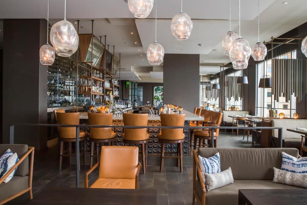 A modern hotel bar and lounge at the Four Seasons Hotel Seattle featuring a row of caramel leather stools along a marble bar, clustered blown-glass pendant lights overhead, gray upholstered lounge seating with patterned throw pillows, and large windows with a city view in Seattle, Washington, USA.