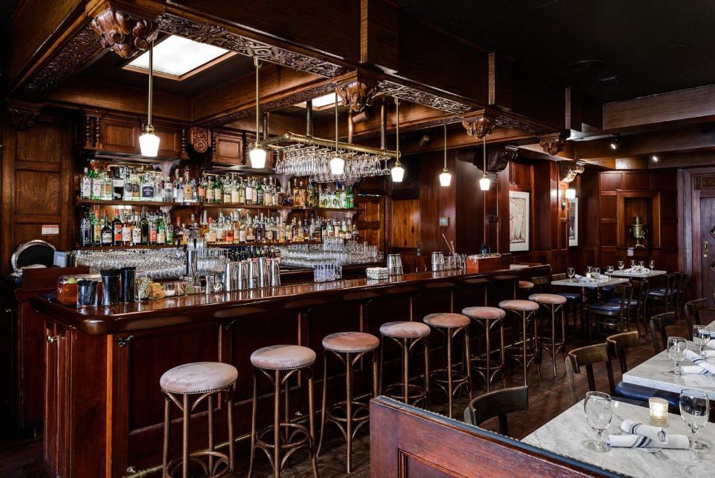 A historic hotel bar at the Hotel Sorrento featuring an ornately carved dark wood bar back stocked with liquor bottles, a wine glass rack overhead, pink velvet stools along the bar, pendant lighting, and marble-top dining tables set with wine glasses in Seattle, Washington, USA.